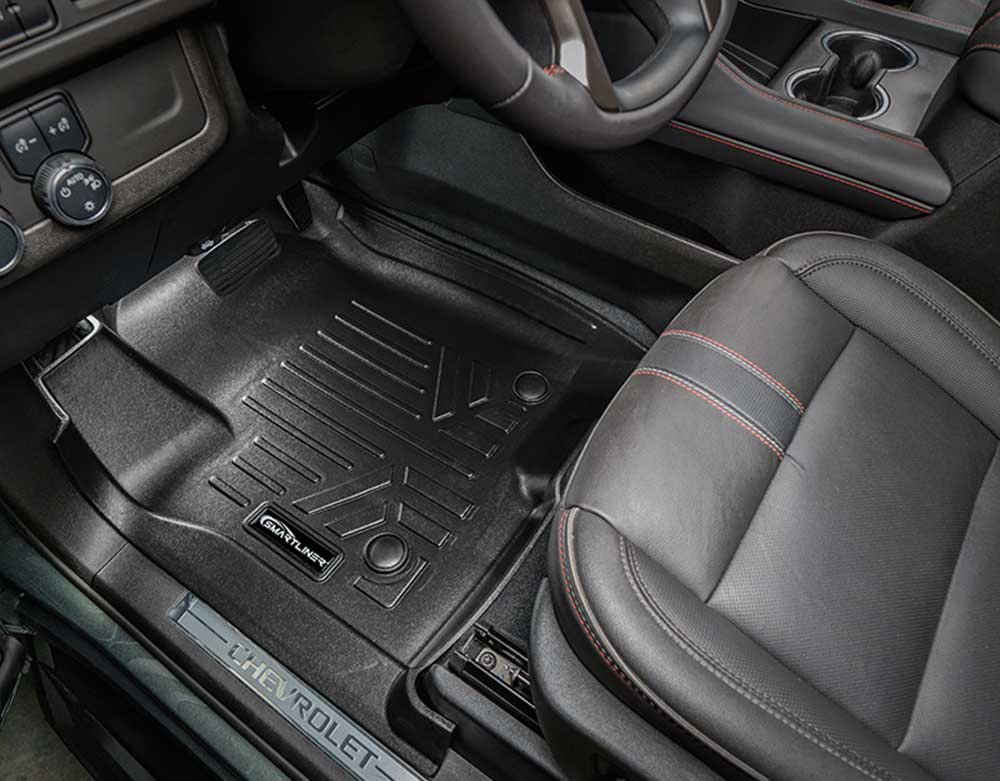 Black rubber car floor mats installed in a vehicle interior, shown with a black leather seat and dashboard. A small Chevrolet logo badge is visible on one mat, and a Chevrolet nameplate is visible on the door sill.
