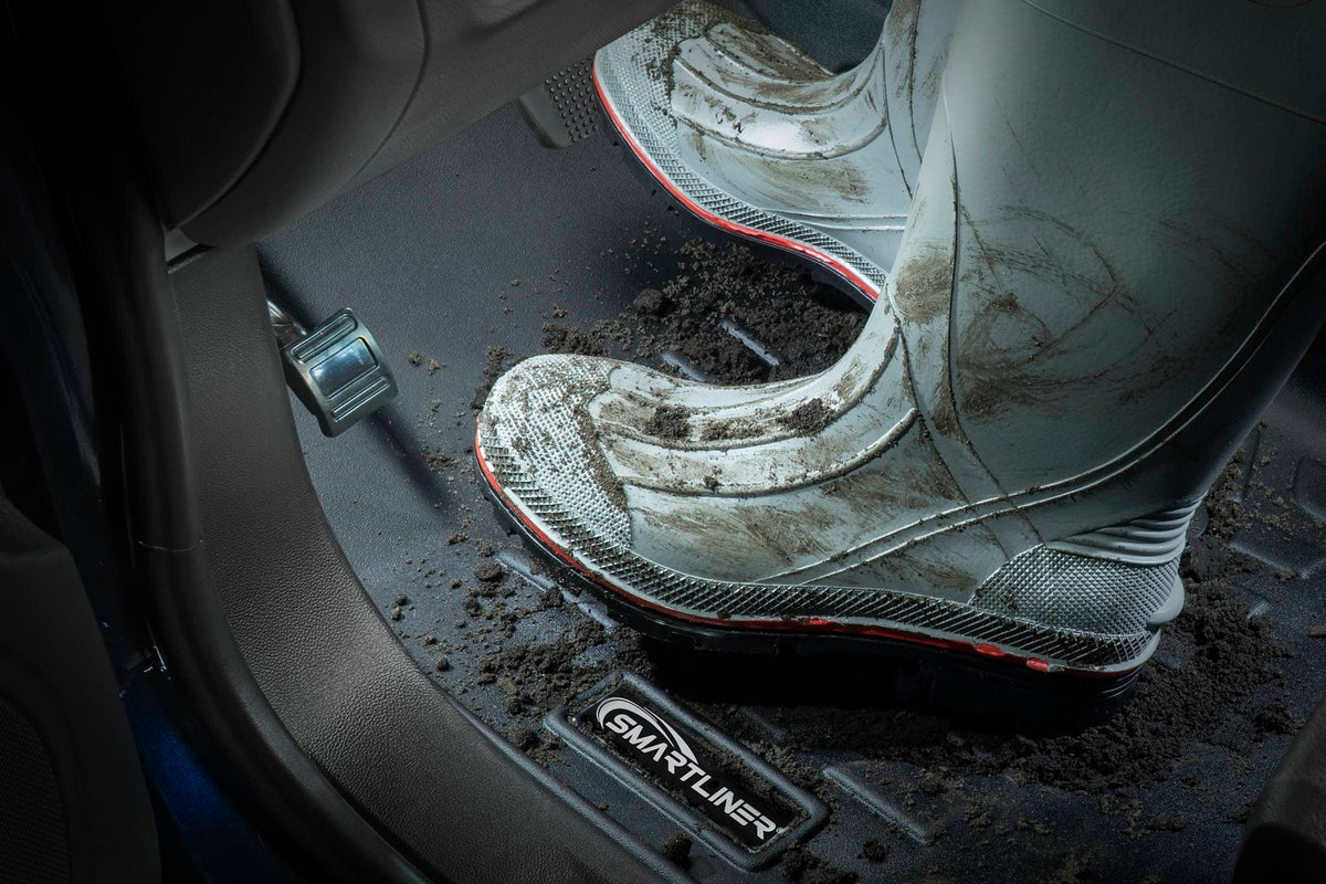 A close-up of a person's muddy gray and white rain boots standing in dark, dirty car footwells next to a black Smartfitter floor mat.